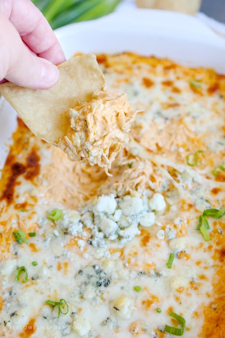 Una mano con un chip de tortilla para obtener una cucharada de salsa de pollo de búfalo de un plato de cazuela