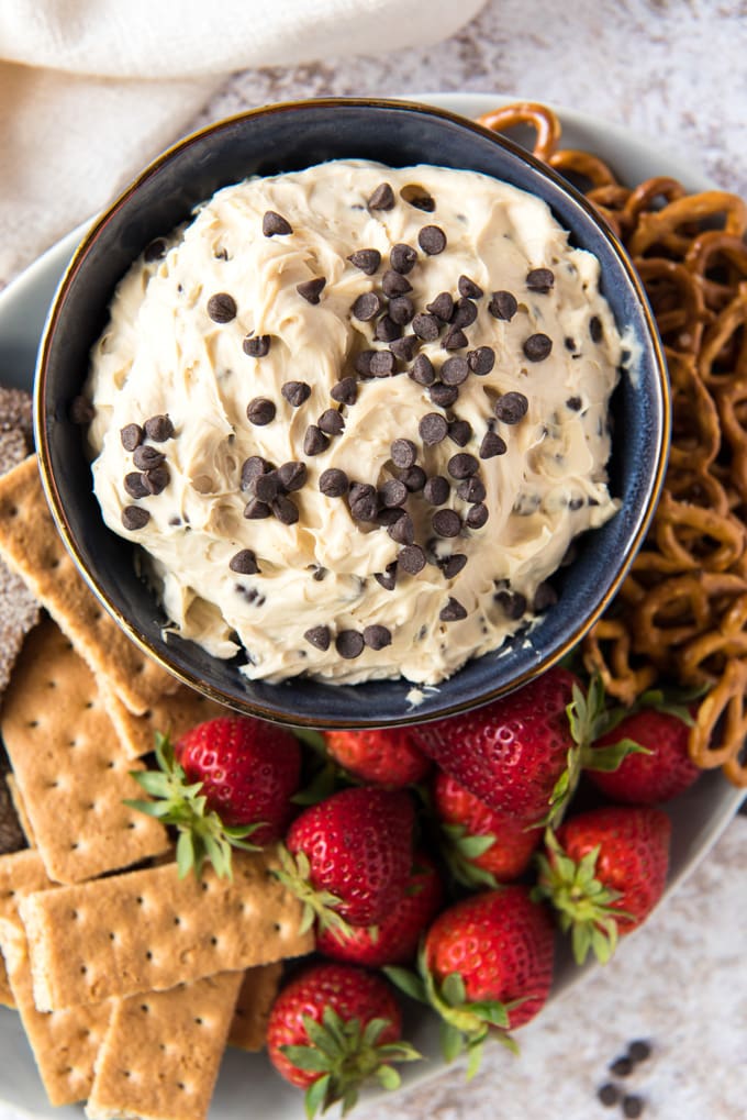 a white plate with graham crackers, strawberries, pretzels, a small blue bowl with cookie dough dip