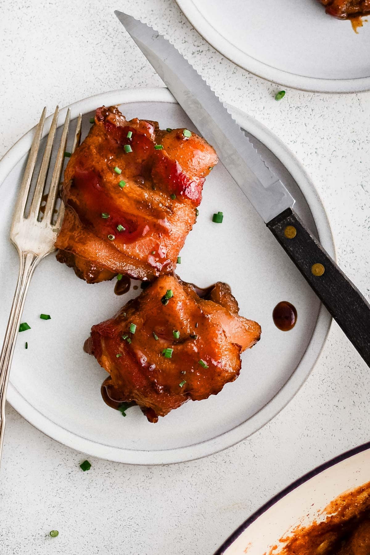 Two chicken thighs wrapped in a crispy bacon on a white plate with a fork and knife.