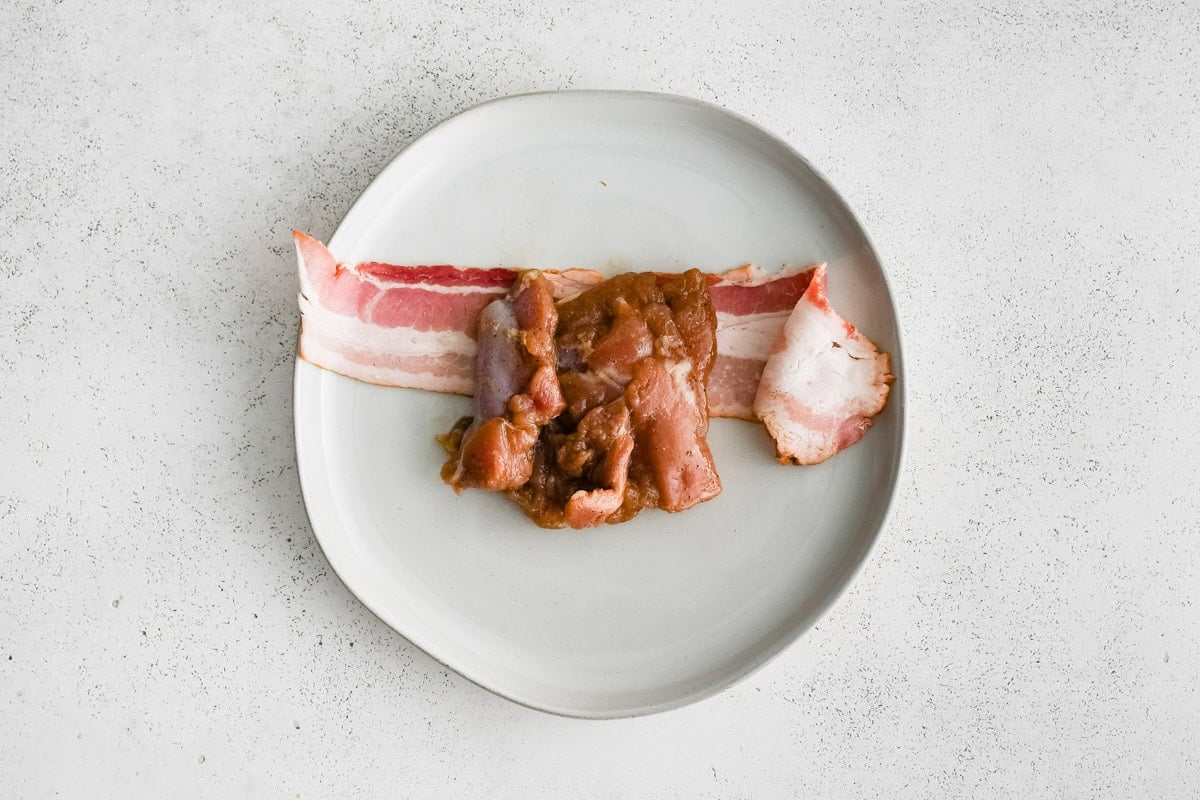Chicken thigh and a piece of raw bacon on a white plate.
