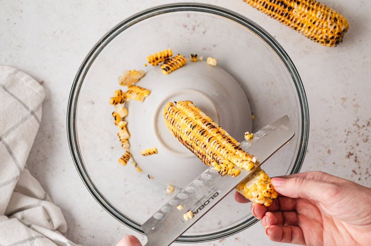 Corn on the cob held by a hand with a knife slicing off the kernels into a bowl.