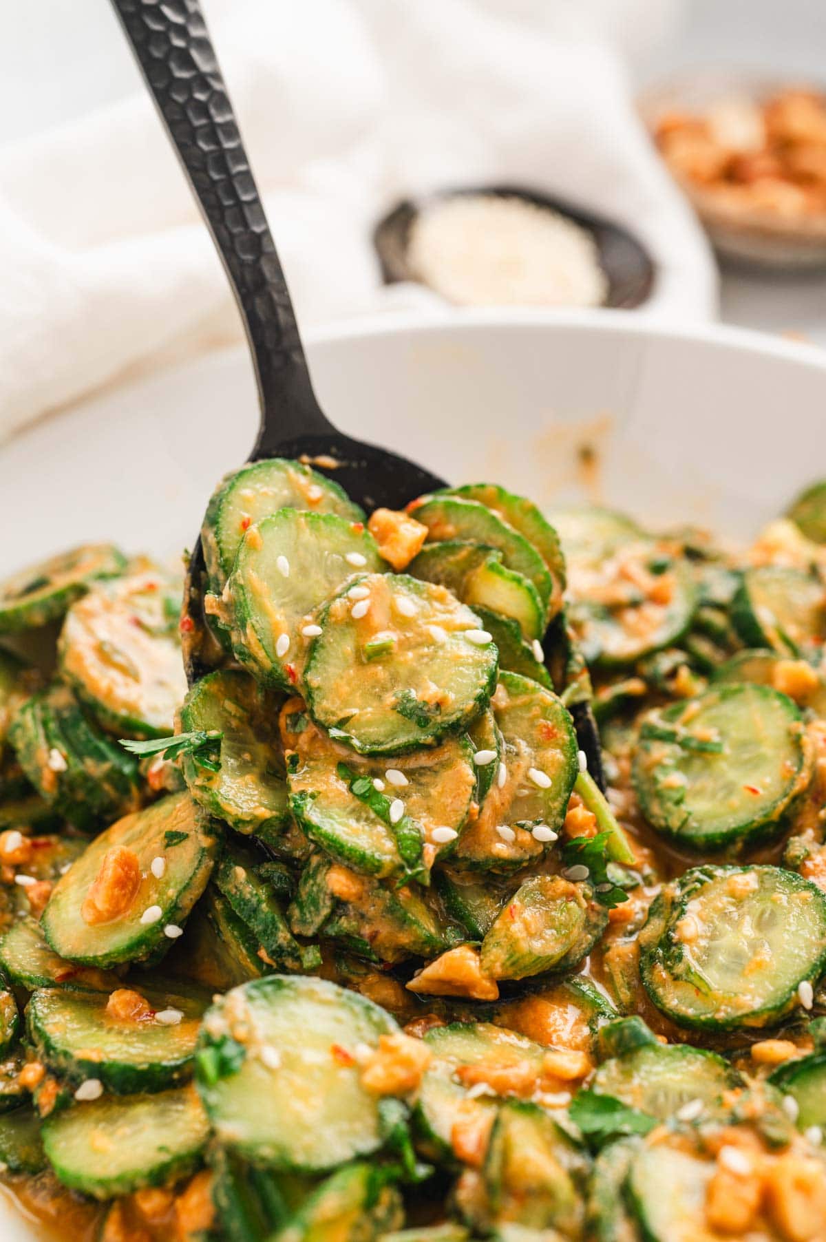 Sliced cucumber salad with peanut dressing in a white bowl with a black serving spoon.