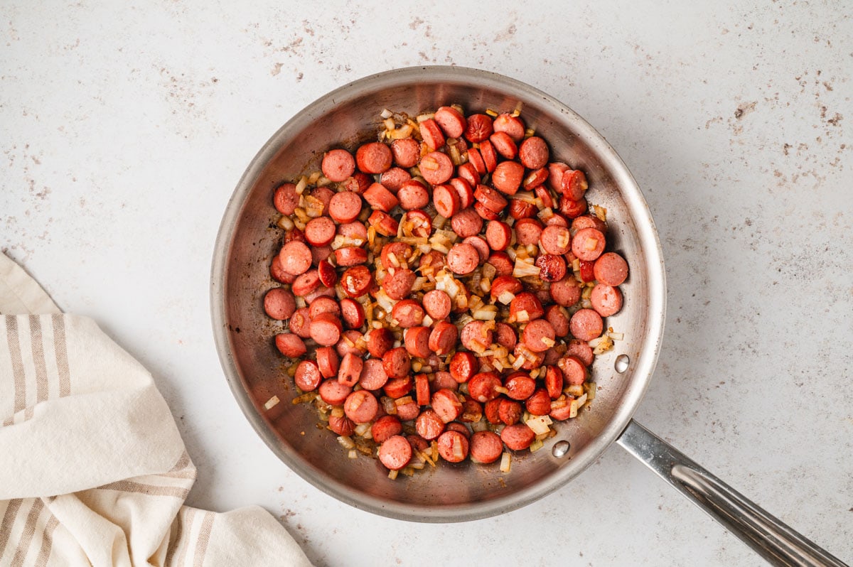 Dice onions and sliced hot dogs frying in a skillet.