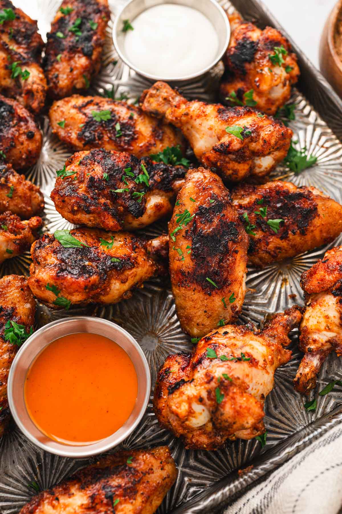 Grilled chicken wings on a baking tray with 2 dishes of buffalo sauce and ranch.