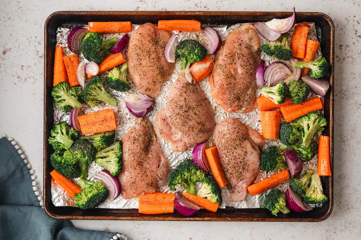 Uncooked chicken and broccoli and carrots on a sheet pan.