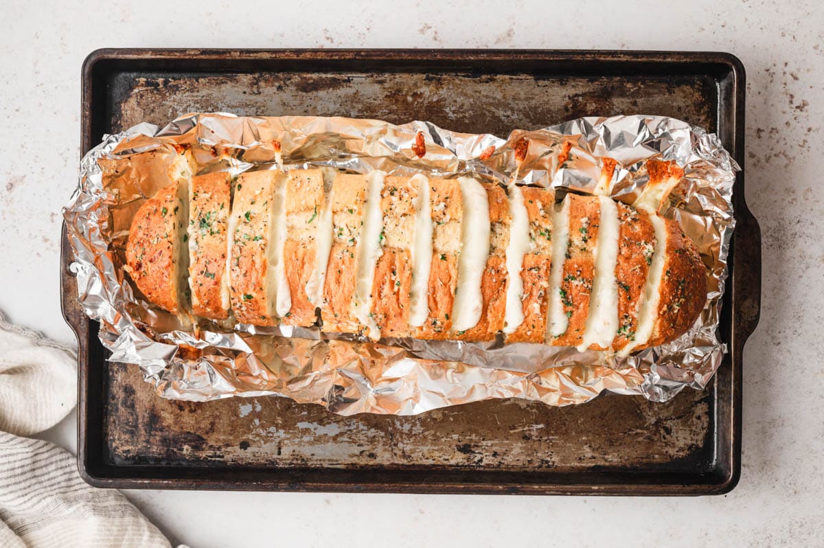 Sliced italian loaf with garlic butter and Mozzarella between the slices sitting on foil on a baking sheet.