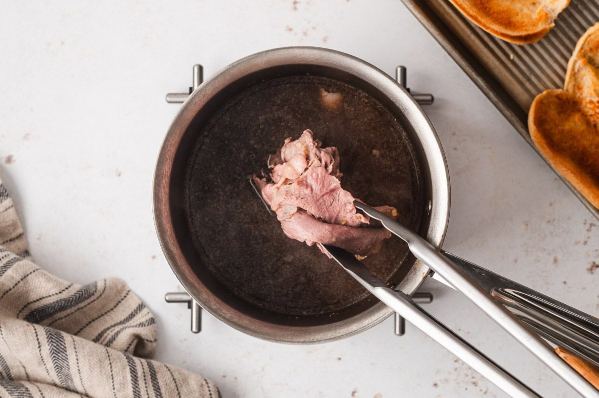 Roast beef with tongs held over a pot of beef stock.