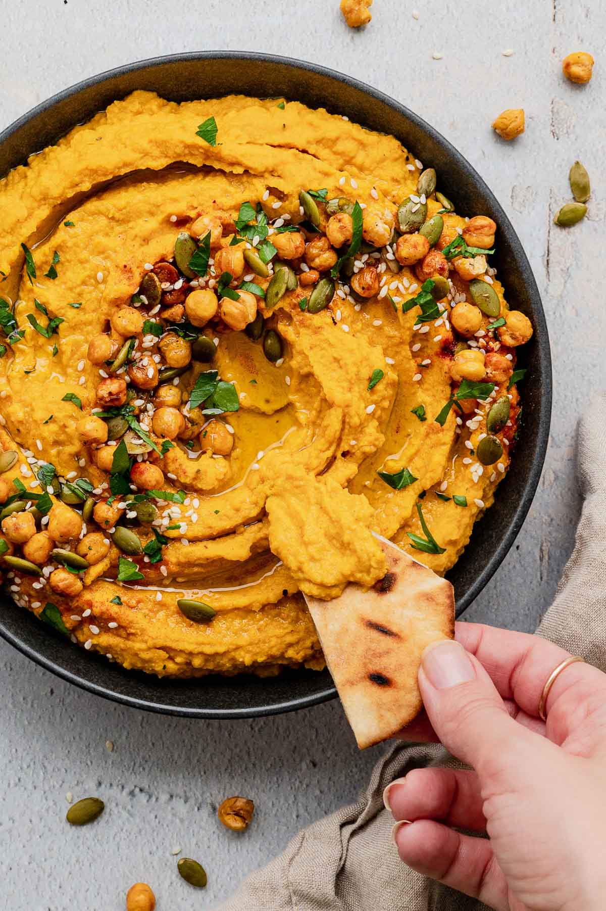 Pumpkin hummus topped with chickpeas, parsley and sesame seeds, a hand holds a pita triangle.