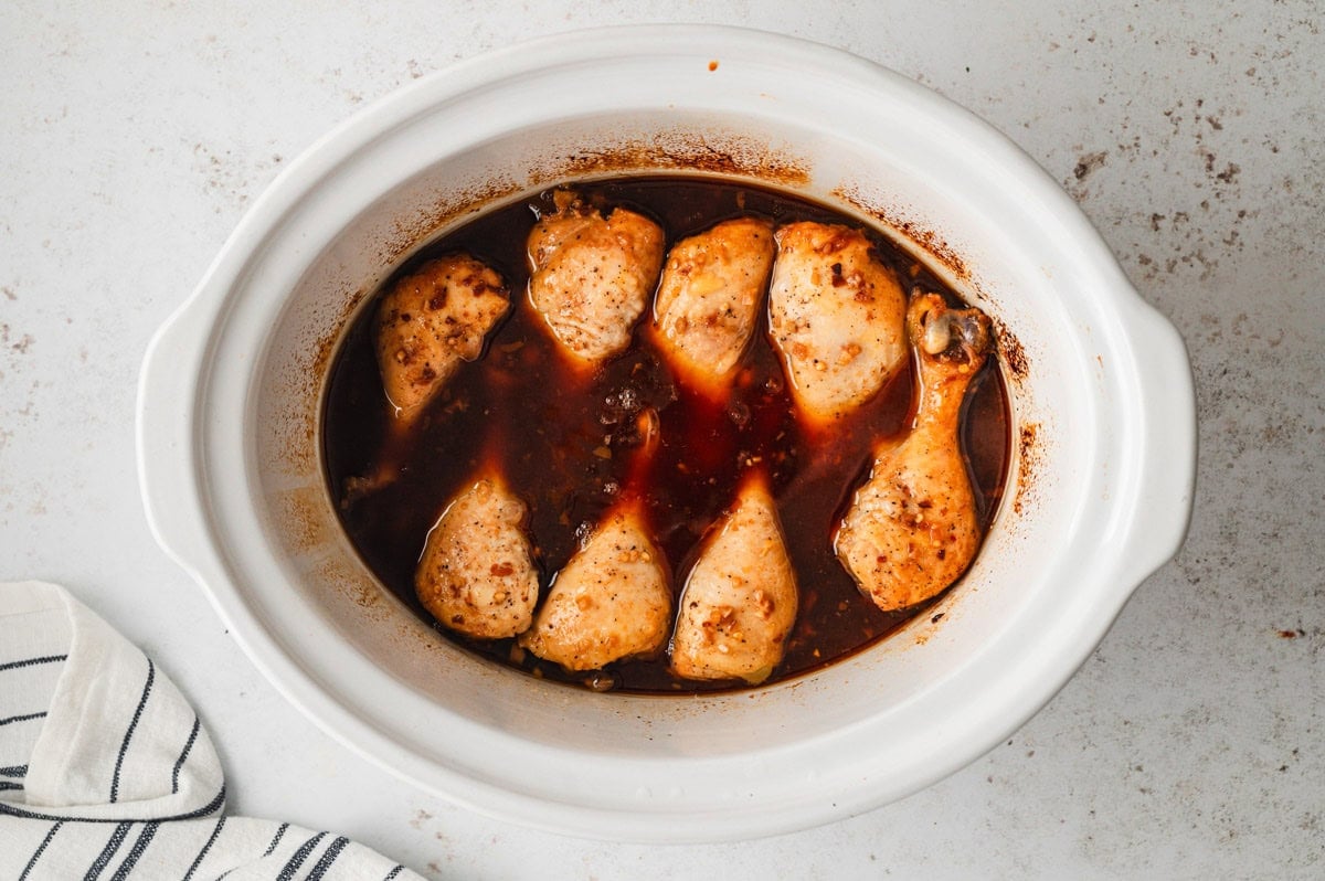 Chicken drumsticks with brown sauce in a white slow cooker.