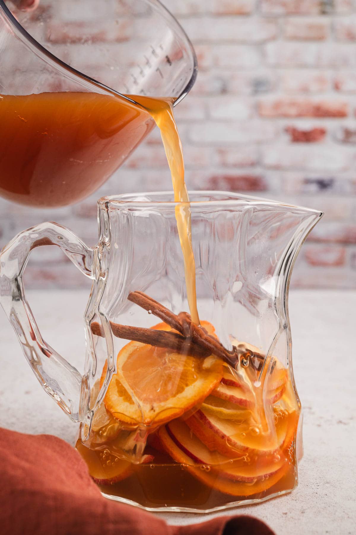 Glass pitcher with cinnamon sticks and orange slices, with apple cider being poured in.