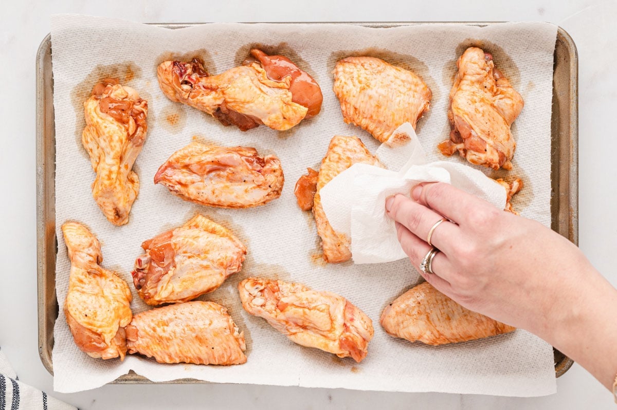 chicken wings on a baking tray with a hand patting dry with paper towel.