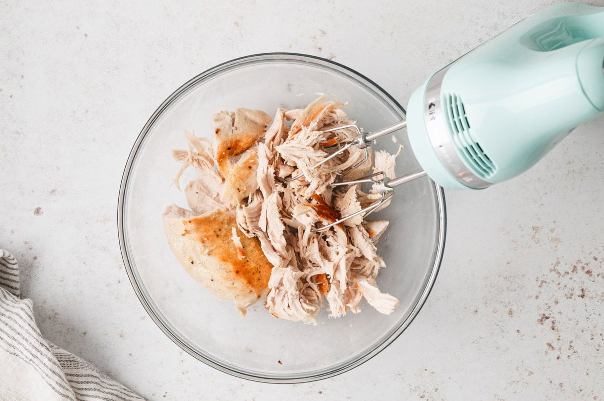 Hand mixer in a bowl with shredded chicken.