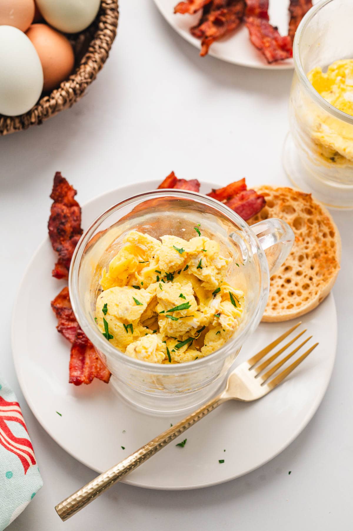 Glass mug with scambled egg on a plate with bacon and english muffin.