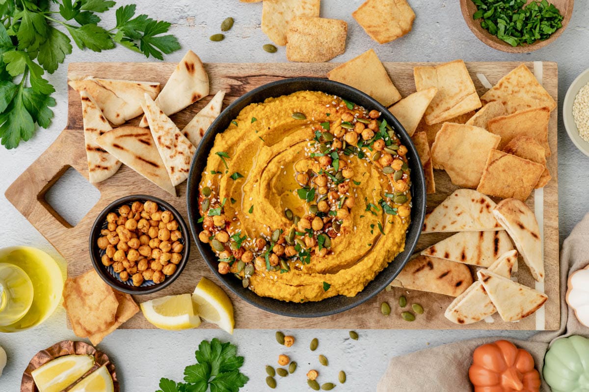 Pumpkin hummus in a bowl, surrounded by pita chips and garnishes.